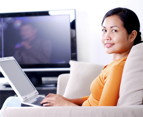 Woman seated on a couch using a laptop, with TV on in the background