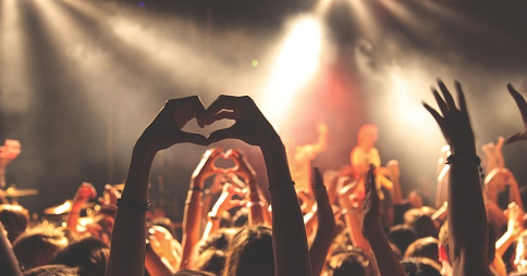 Audience cheering whilst facing the stage at a live performance. A person's hands are raised in the foreground making a 'heart' shape.