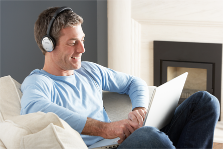 Man on sitting on a couch, smiling whilst wearing headphones connected to a laptop