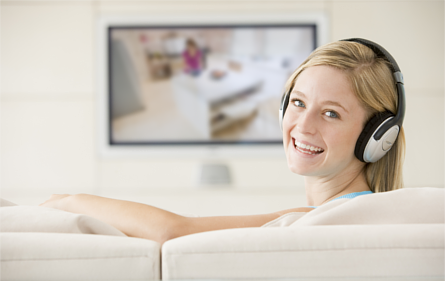Woman wearing headphones while watching TV. She is sitting on a couch, looking over her left shoulder and smiling.
