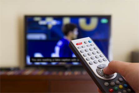 Right hand holding remote control in front of captioned sporting event on television