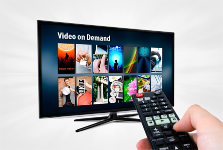 Left hand pointing a remote control at a Smart TV displaying a Video on Demand app
