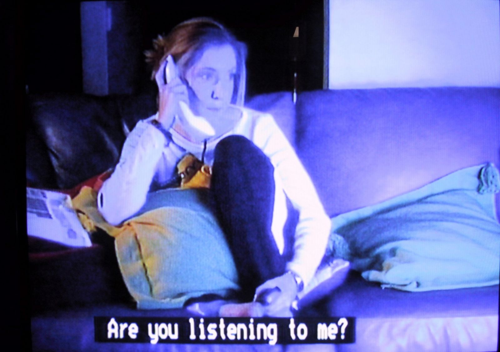 Example of a standard caption: a frame from a movie showing a girl on the phone watching a television with the caption below 'Are you listening to me?'