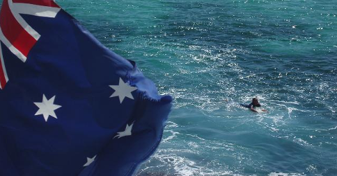 Australian flag waving in the foreground; in the background, a surfer swims in the water off the New South Wales coast