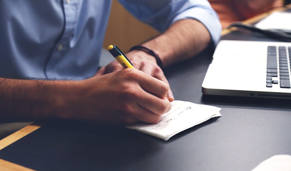 A close up of a man writing on paper on a desk with laptop next to him